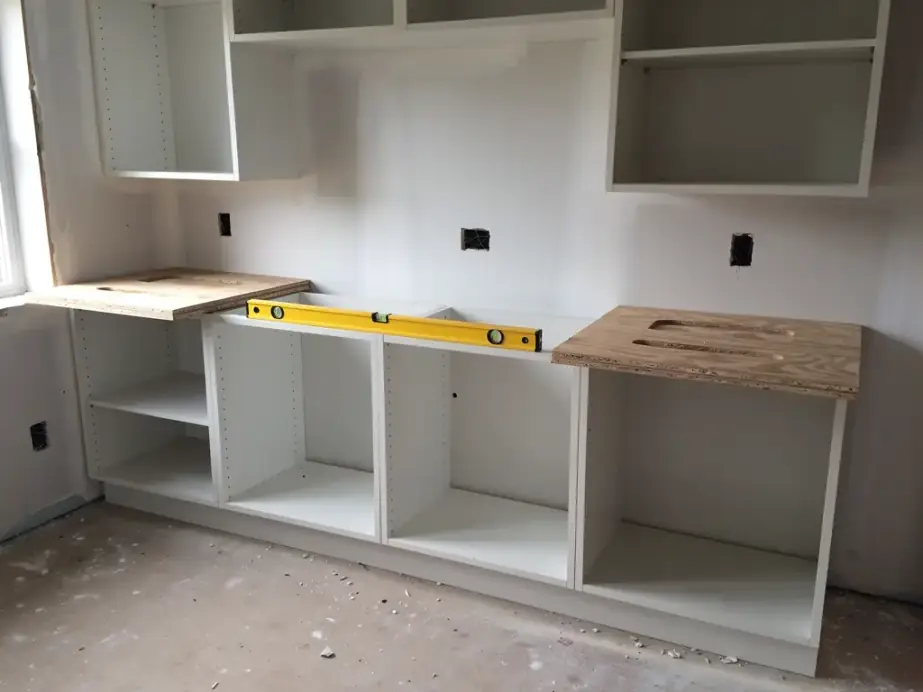 Kitchen cabinet boxes set on a primed wall with no doors fitted yet and a level resting on the base run